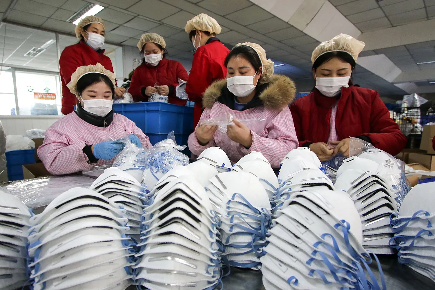 Six workers wearing face masks and hair nets produce protective facemasks, seen in front of them in stacks, at a factory.