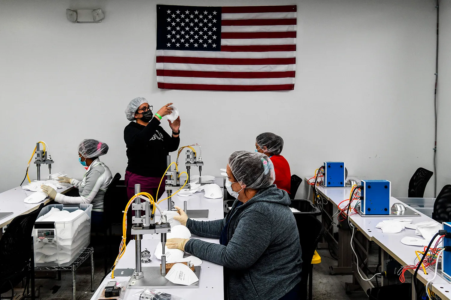 Workers wearing masks and hairnets sit at tables, with one standing, as they make protective face masks. Above them on the wall is a U.S. flag.