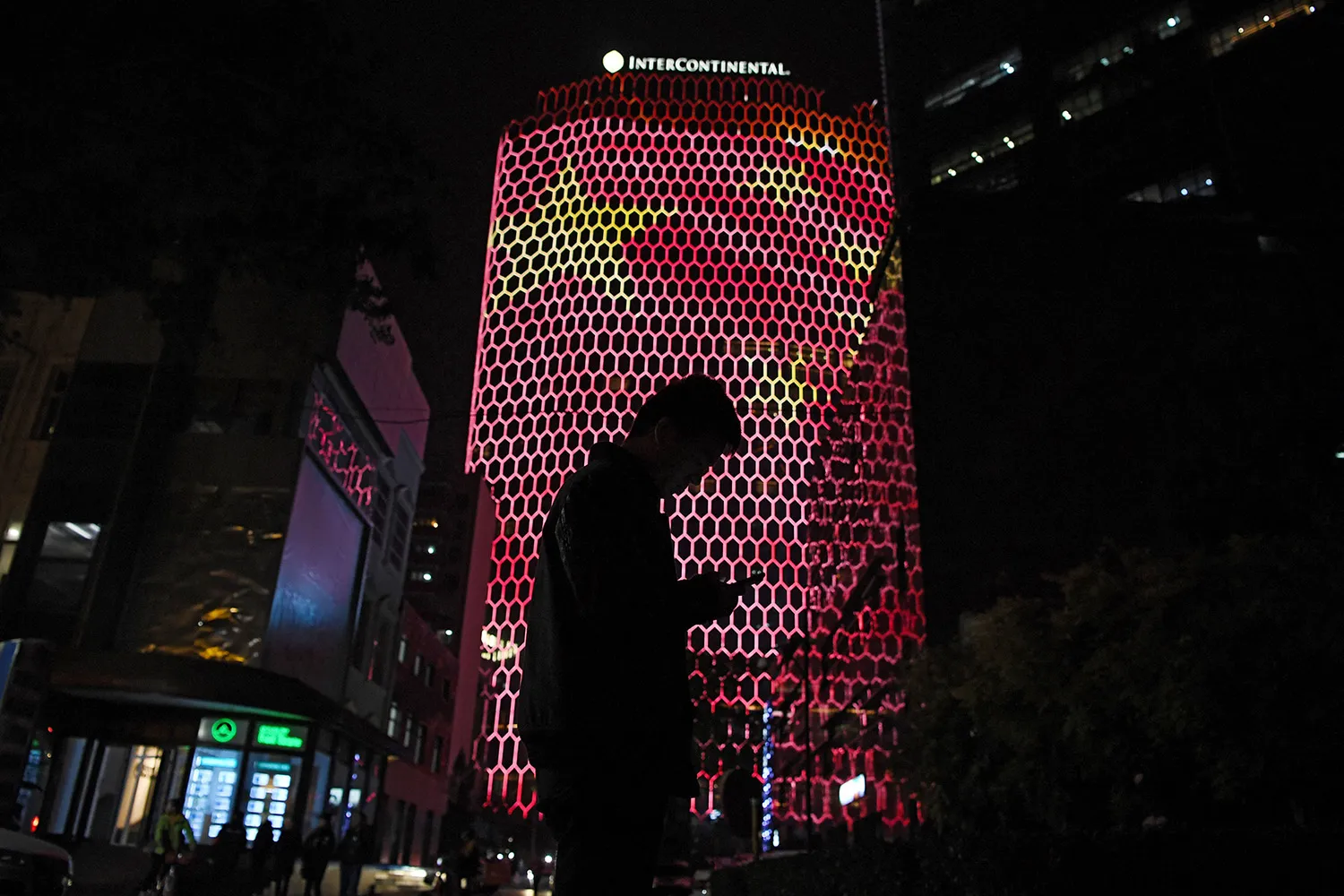 A man looking at his phone is silhouetted against a building with a net-like, tech texture overlaid with the Chinese national flag. A sign atop the building says: "Intercontinental."