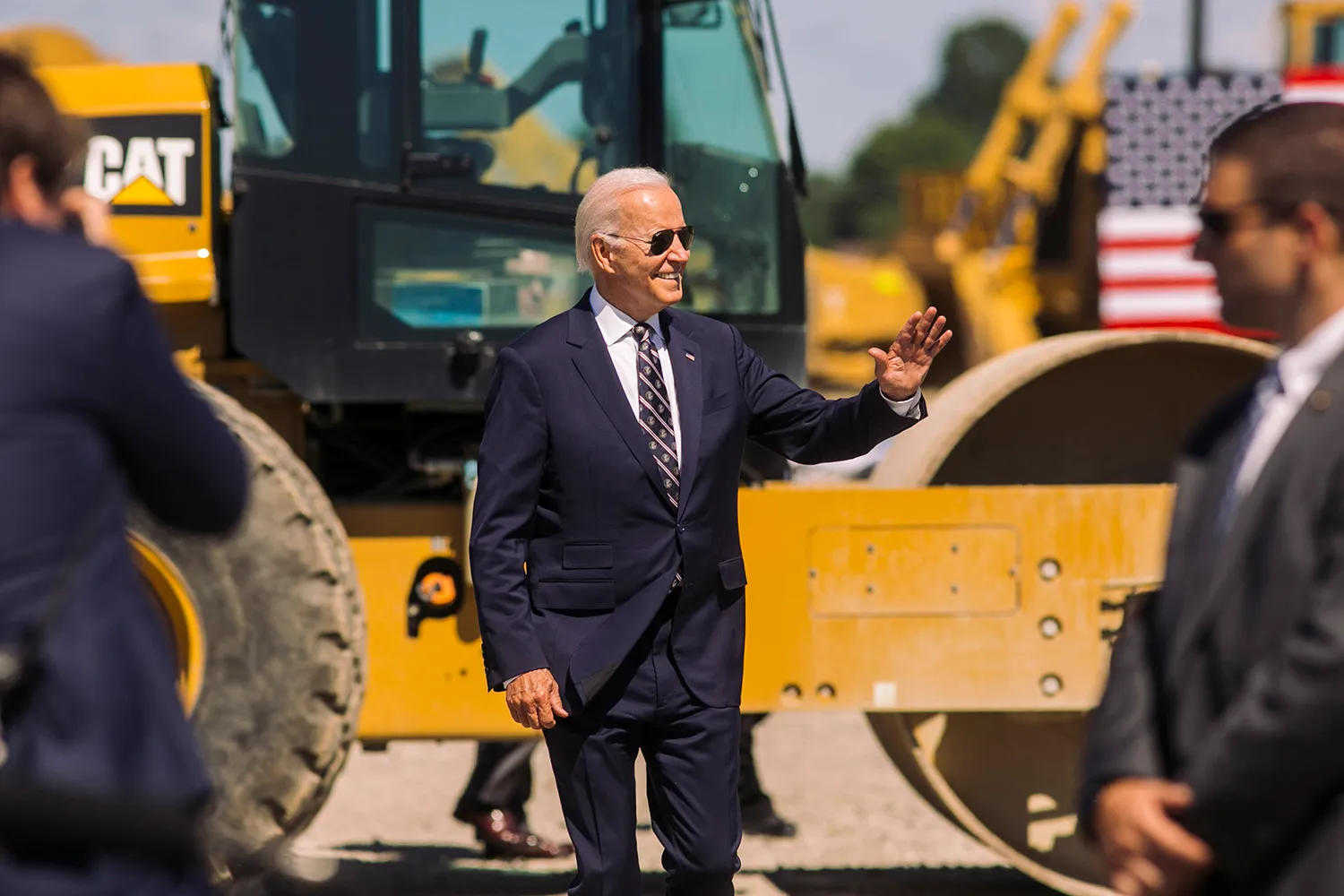 U.S. President Joe Biden, wearing a suit and tie and sunglasses, smiles as he waves in front of a heavy machinery outside a factory. A secret service agent is seen out of focus at right as is a U.S. flag behind him.