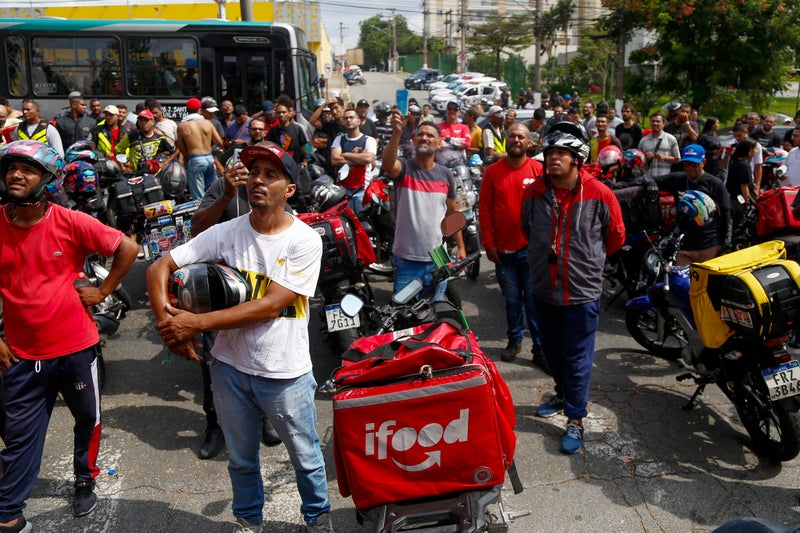 A group of delivery riders, wearing helmets and colorful uniforms, gather in a street, some holding helmets and others looking towards the sky, while motorcycles with delivery bags marked 'ifood' are lined up in the background.