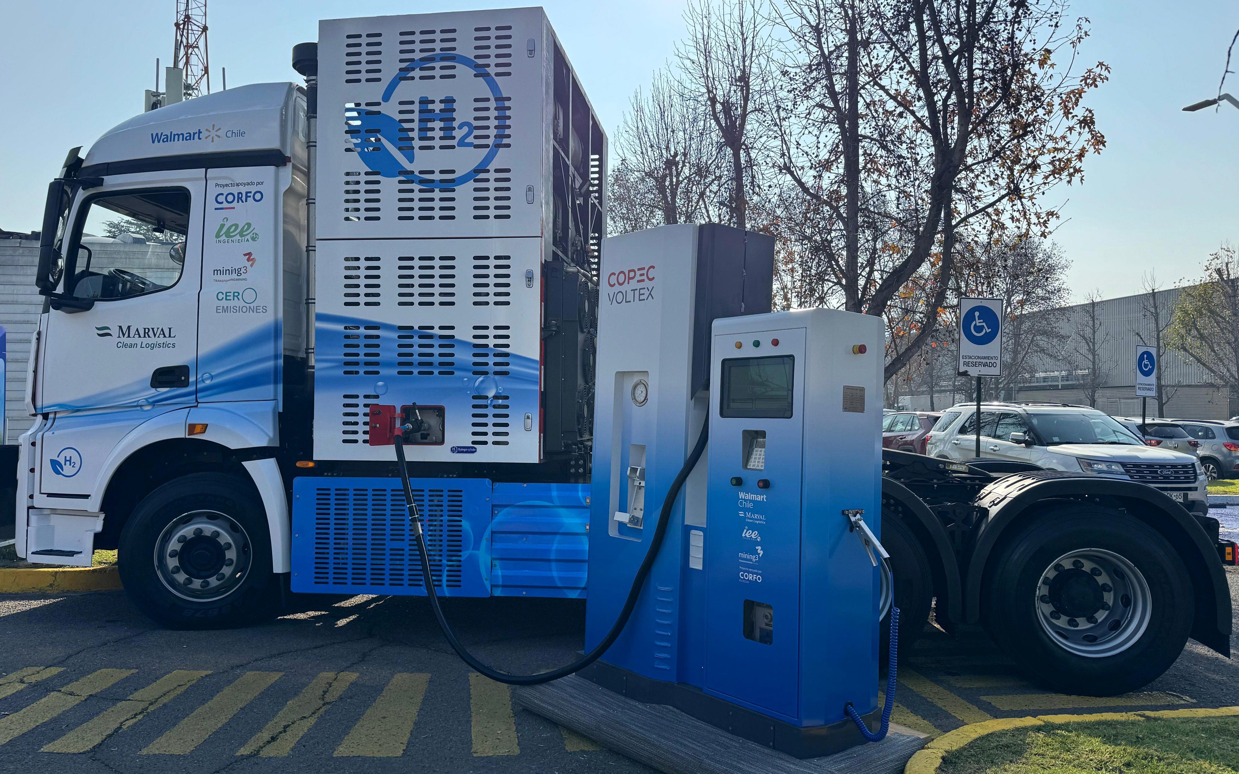 A white and blue truck cab that says Walmart Chile as well as showcasing many other logos charges at a hydrogen station.
